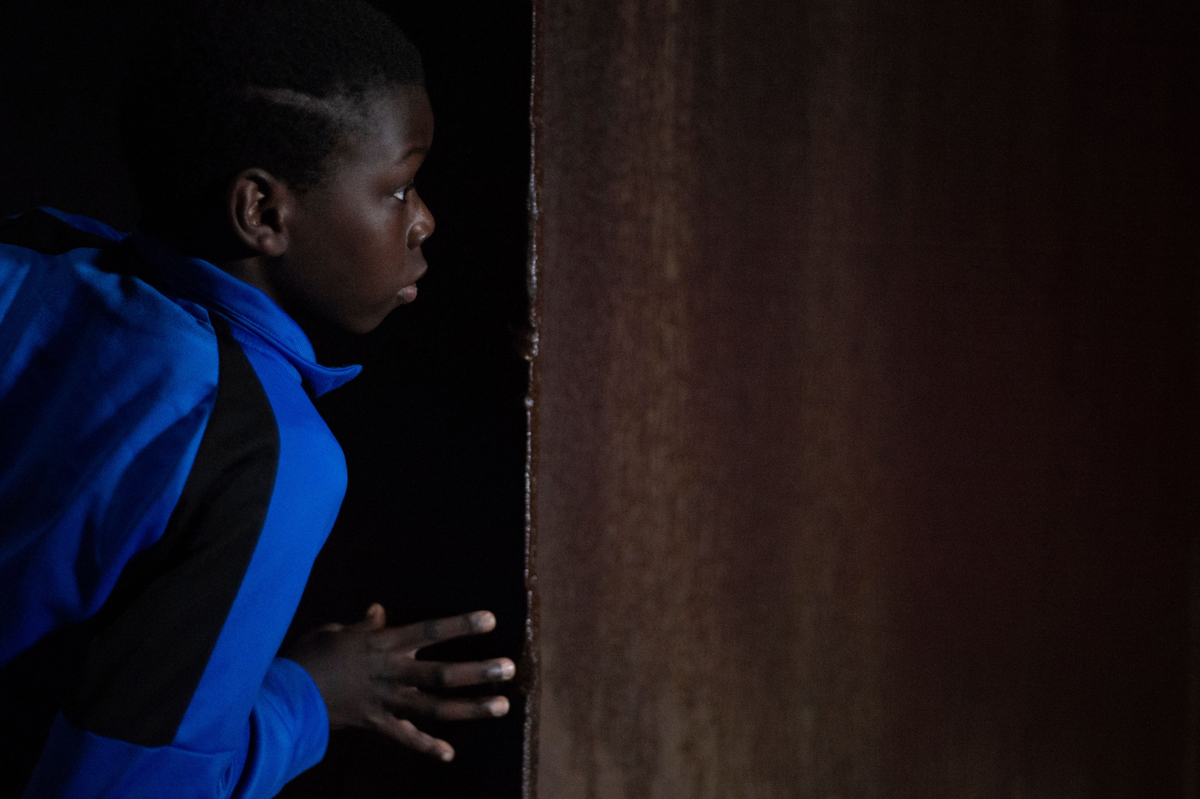 Young Black boy in blue jacket peers around a dark corner