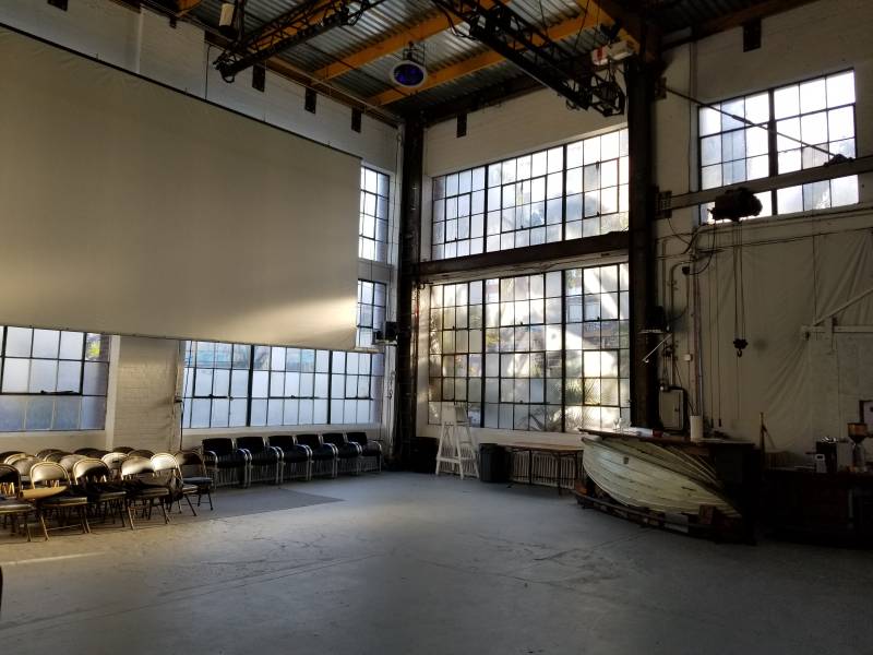 a large room or performance space with folding chairs, high ceilings, and a wall full of windows with light streaming in