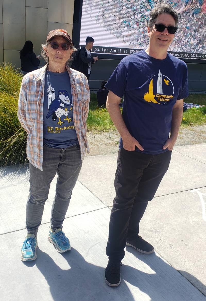 A senior woman and a middle-aged man stand near a grassy area on a street corner. They are both wearing t-shirts featuring falcons.