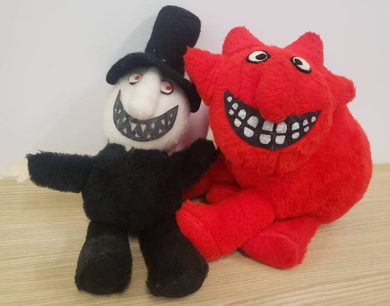Two stuffed toys. One is a white-faced figure with red eyes and grin of fangs, wearing a black suit and top hat. The other is a grinning red devil with pointed tail.