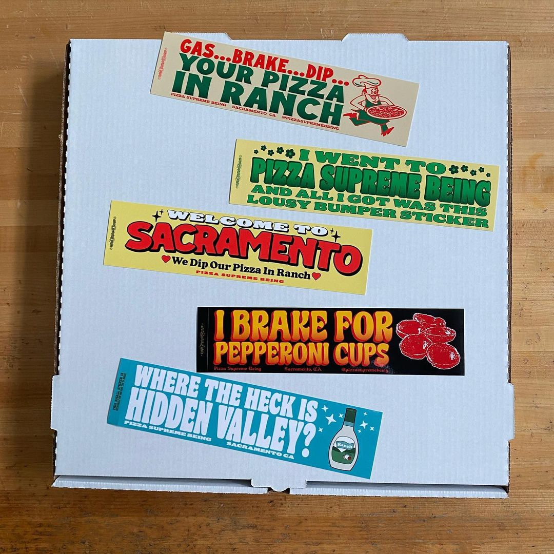 Bumper stickers arranged on a white pizza box: "Gas...Brake...Dip...Your Pizza in Ranch," "I Went to Pizza Supreme Being and All I Got Was This Lousy Bumper Sticker," "Welcome to Sacramento, We Dip Our Pizza in Ranch," "I Brake for Pepperoni Cups," and "Where the Heck Is Hidden Valley?"