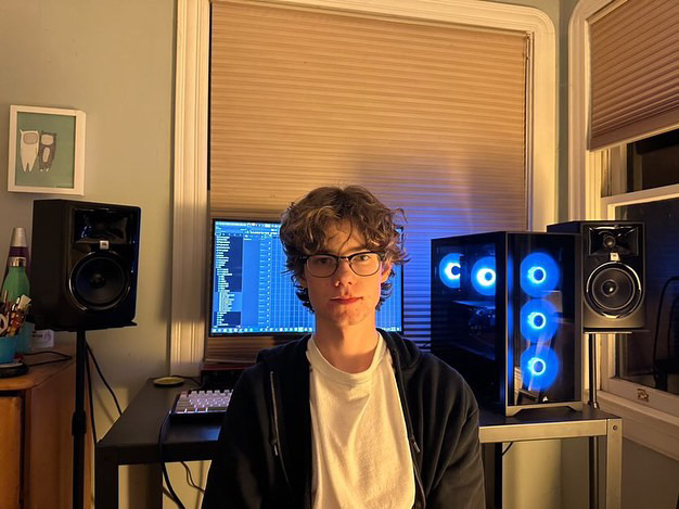 A young man sits in his room in front of a computer and speakers.