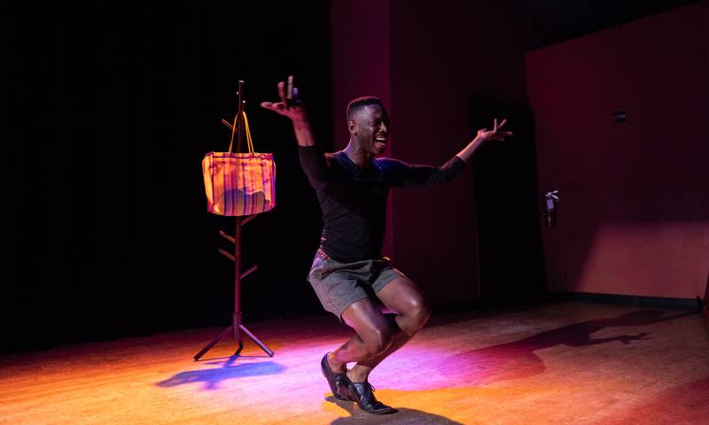 a young Black man wearing shorts and a long-sleeve shirt moves with his arms in the air on a pink-lit stage during a performance