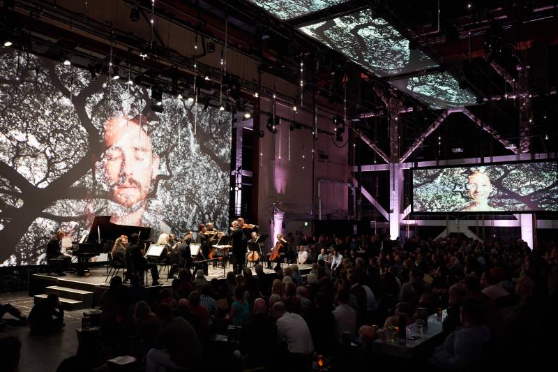 a crowd of people sits watching classical musicians perform in a dark club-like space with large artworks projected onto the walls and ceiling