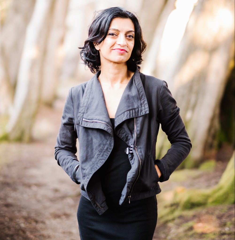 Woman with shoulder length black hair stands with hands in jacket pockets with trail and trees behind her