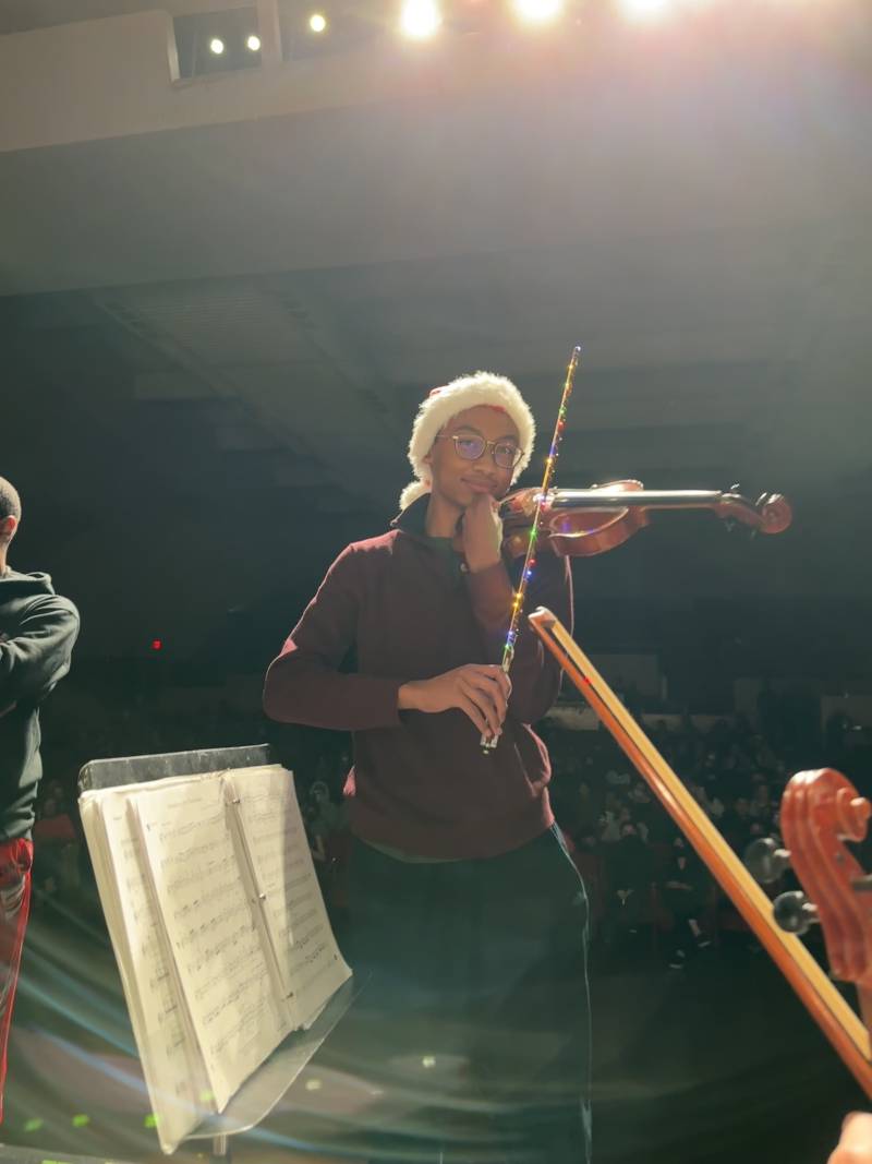 A young man stands on stage holding his violin and wearing a Santa hat.