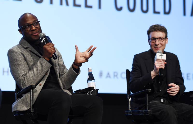 a young Black man with glasses in a grey coat and black pants speaks into a microphone onstage next to a young white man in a black coat and glasses, as part of a panel at a film event