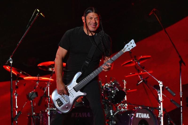A smiling man with long braids, dressed in all black, onstage playing a bass.