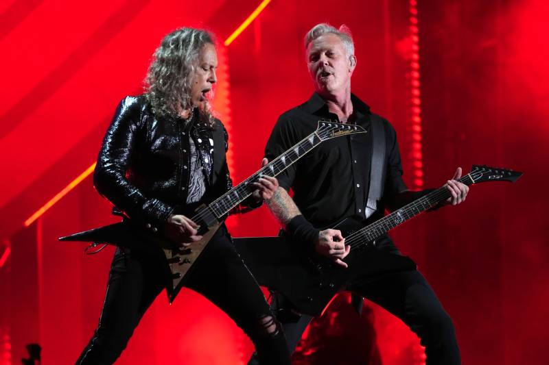 Two rockers stand onstage with wide-legged stances. They are dressed in all black and leather and playing electric guitars. Both have greying hair.