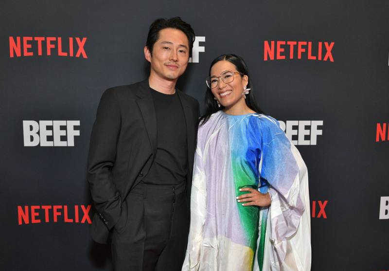 A 30-something Asian man in a black suit and t-shirt stands with a similarly-aged Asian woman wearing a white puffy dress and large spectacles. They are posing in front of a wall with the Netflix logo on.