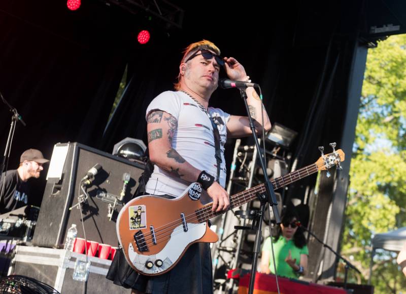 A man wearing a tight white T-shirt, short black skirt and bass guitar, stands on stage peering out from under his sunglasses suspiciously.