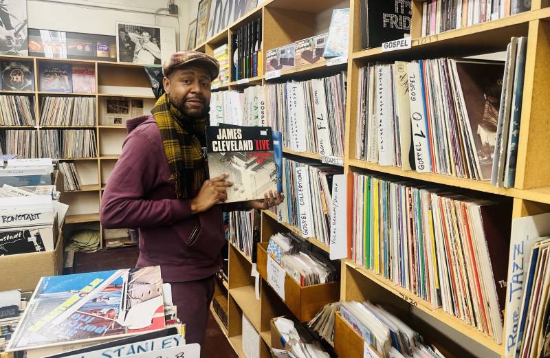 a Black man in a purple jacket holds up a copy of a James Cleveland record while standing in a record store with shelves overflowing with vinyl