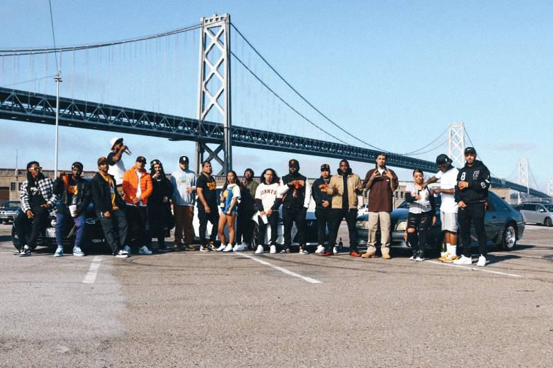A large group of 30 diverse, young hip-hop artists poses in front of the Bay Bridge.
