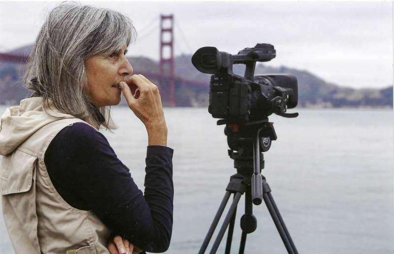 a woman with short gray hair wearing a black long-sleeved shirt and tan vest stands behind a movie camera in front of the Bay and the Golden Gate Bridge