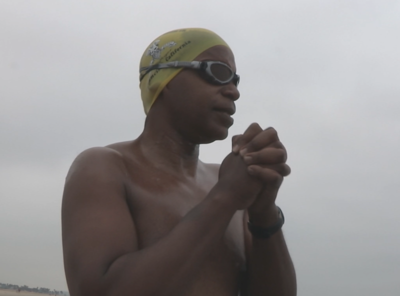 a man with brown skin stands with his shirt off and hands clasped in a yellow swim cap and goggles