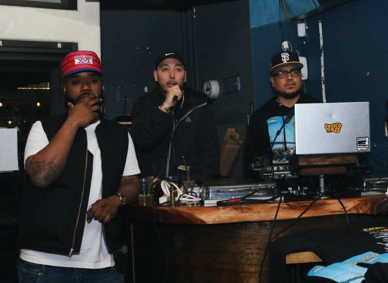 A young Black man and two young Latino men make an announcement behind DJ gear at a party. 