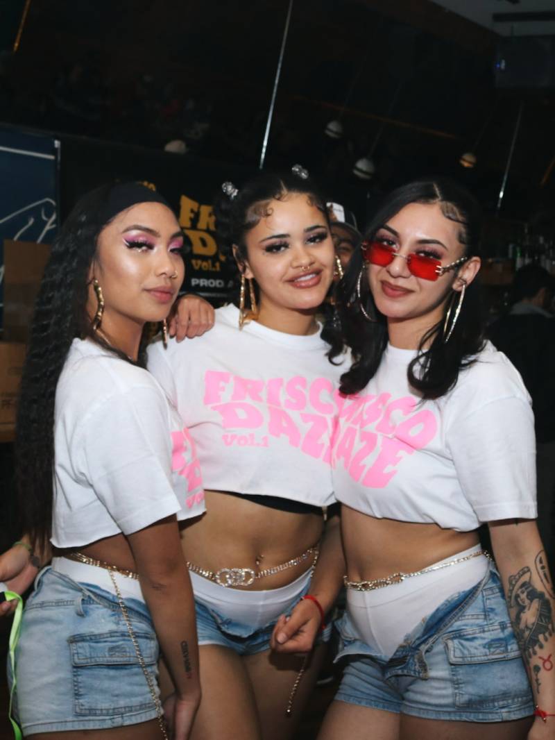 Three young women with matching pink and white Frisco Daze shirts, denim shorts, braids and hoop earrings pose for a photo. 
