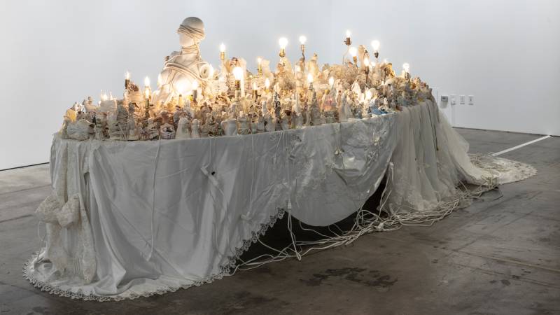 Long table draped in white clothing covered in bare-bulbed lamps and small figurines wrapped in white string