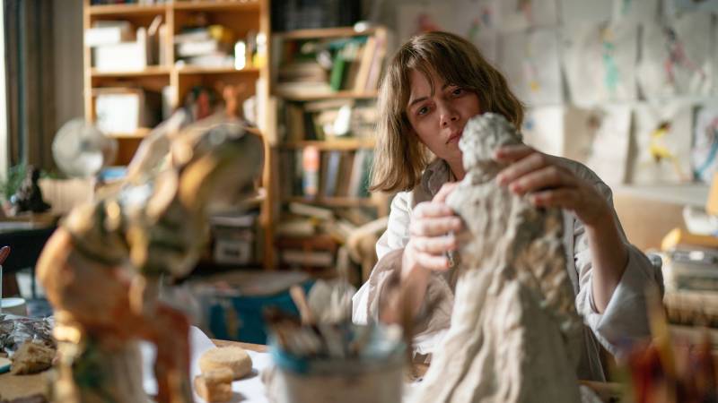 A white woman with a bob works on a clay sculpture of a figure in her book-filled studio