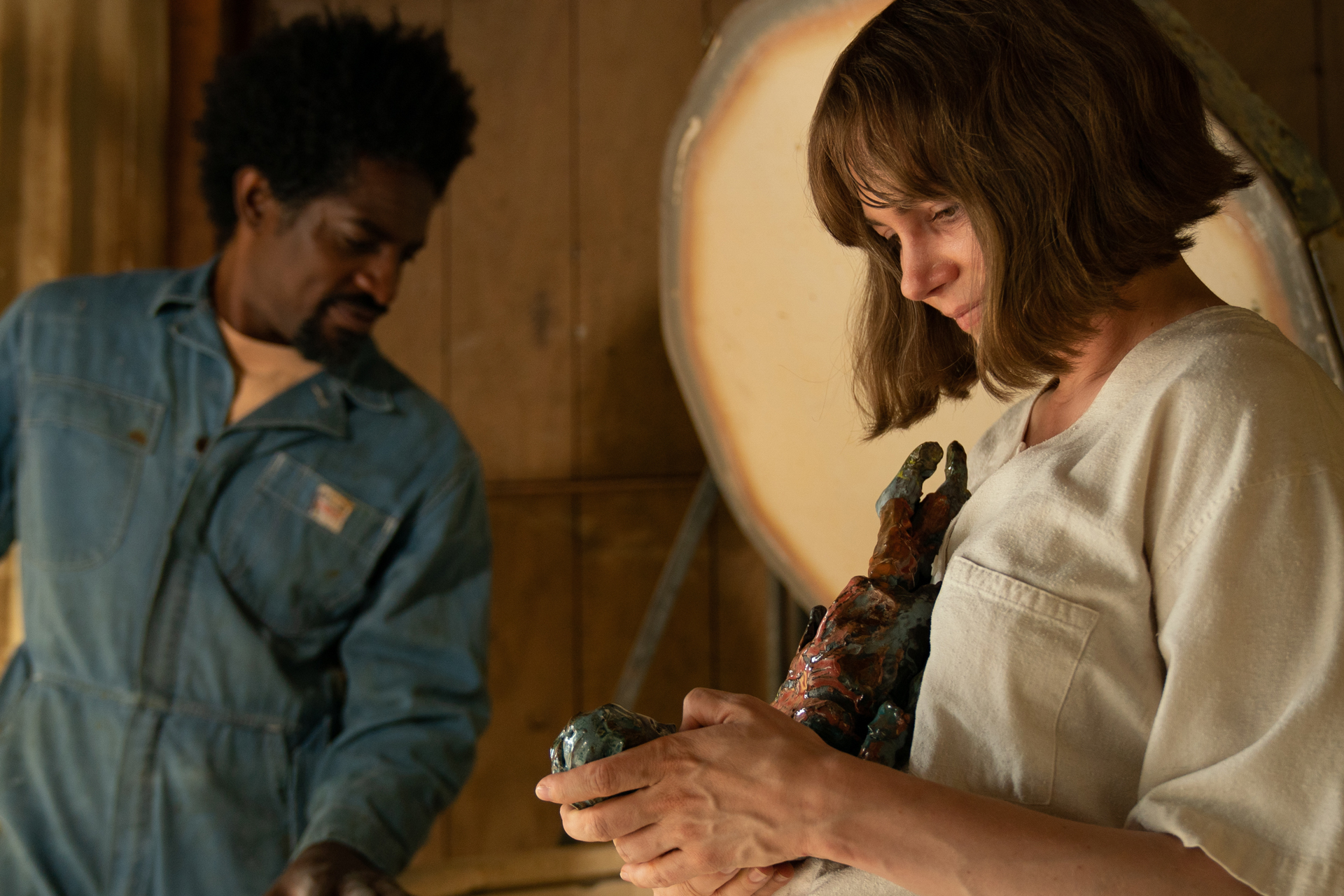 White woman with bob holds sculpture in hands, a Black man in coveralls stands behind her over kiln