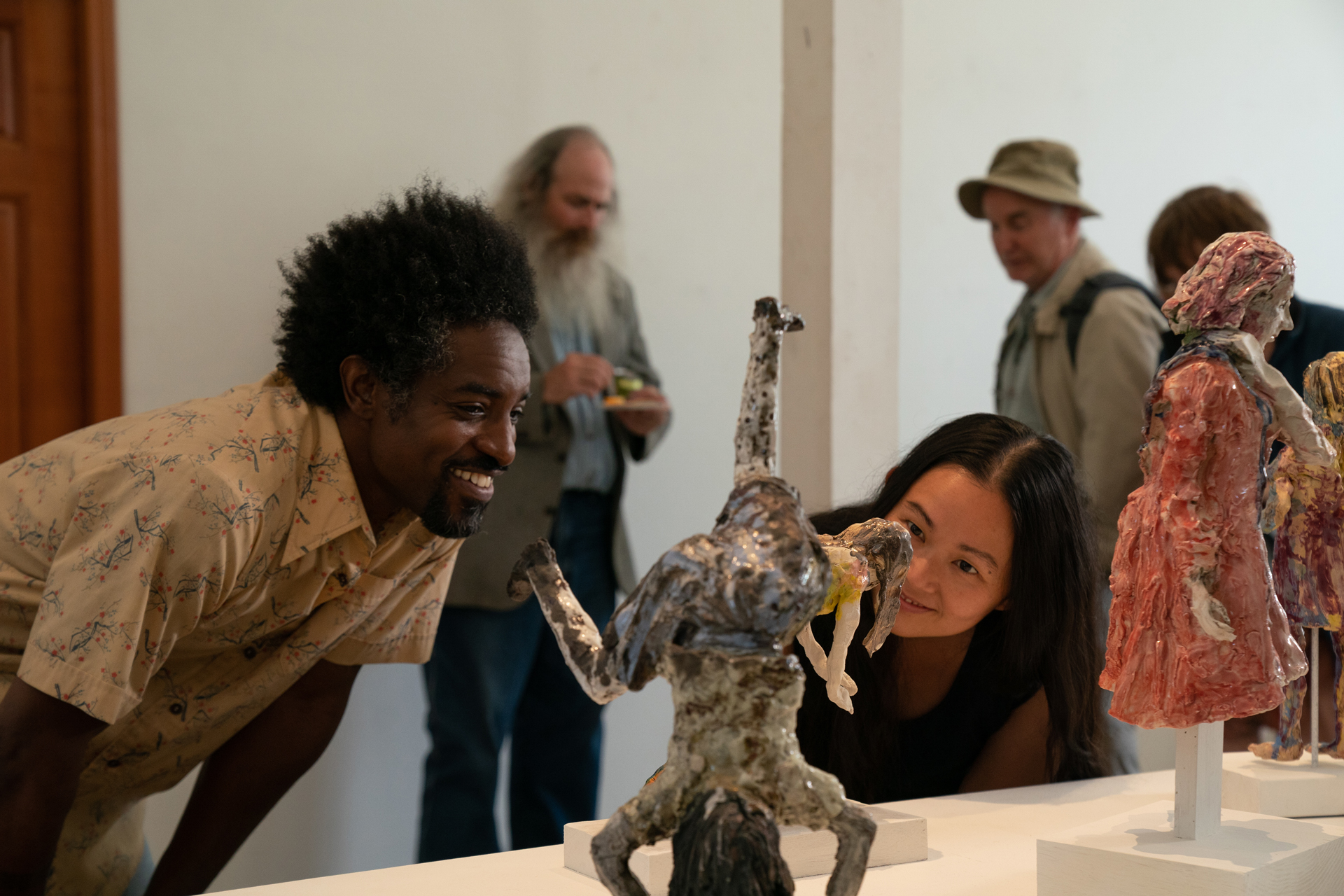 A Black man and an Asian woman lean over to look closely at a small figurative sculpture, smiling