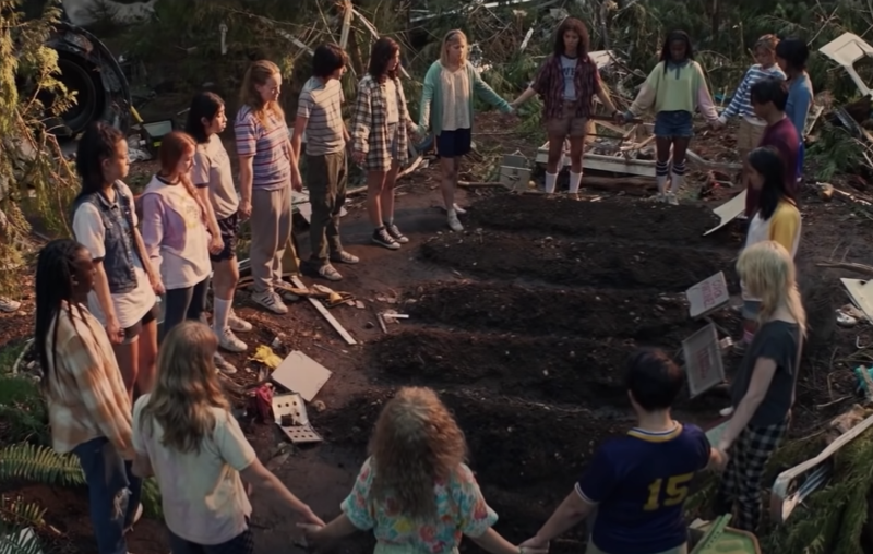 A circle of teens wearing summer clothes gather around five freshly dug graves.