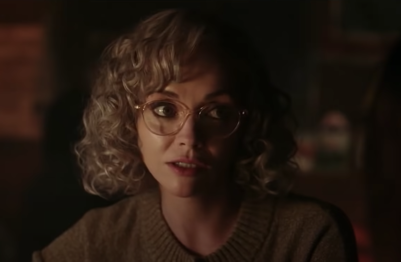 A white woman with a blond curly bob sits in a dark room. She is wearing dowdy spectacles and a sweater.