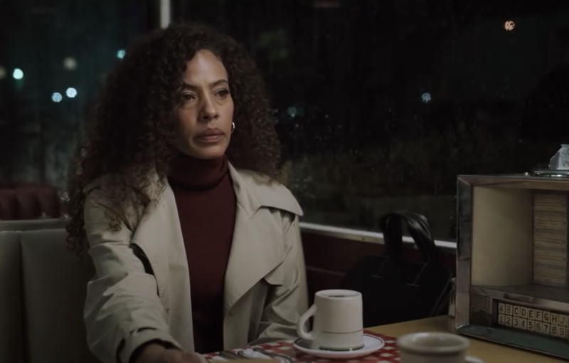 A black woman with long curly hair sits at a diner table, a coffee cup in front of her, a table-top jukebox at her side.