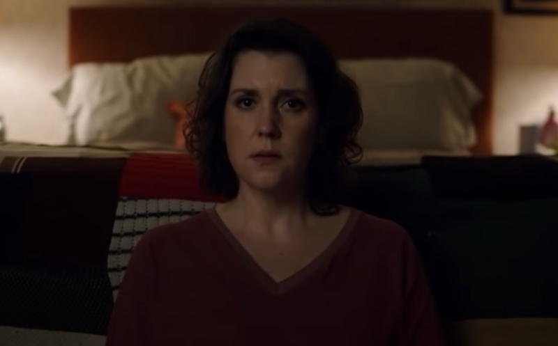 A middle-aged white woman with short dark brown hair sits on the floor, her back leaned against a bed. She looks worried.
