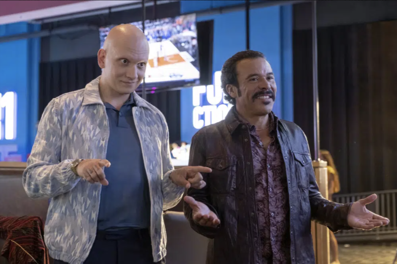 Two men stand side-by-side in a sports bar gesturing to a friend. One is young but entirely bald and wearing a sports jacket. The other is Latino with slicked back hair and mustache and ’70s-style leather jackets.