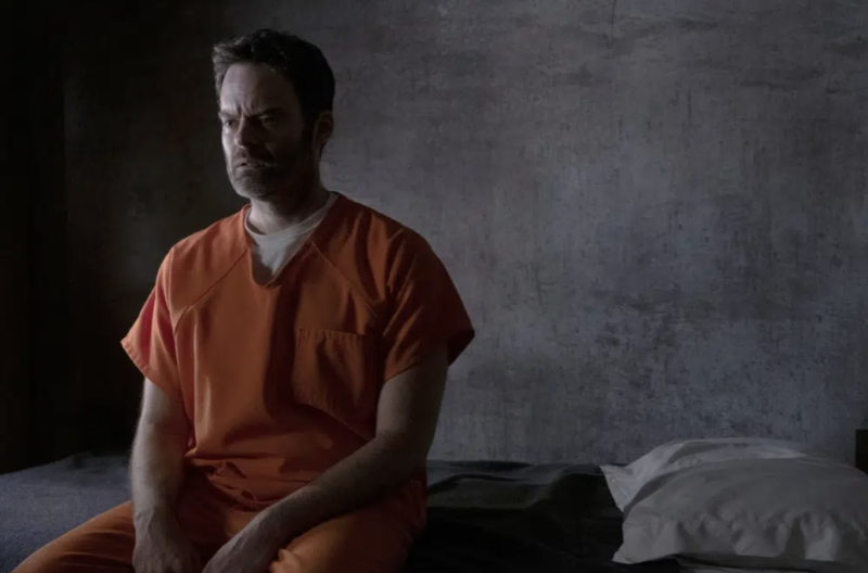 A middle aged man in an orange prison garb sits on the edge of a bed in a prison cell, perplexed expression on his face.