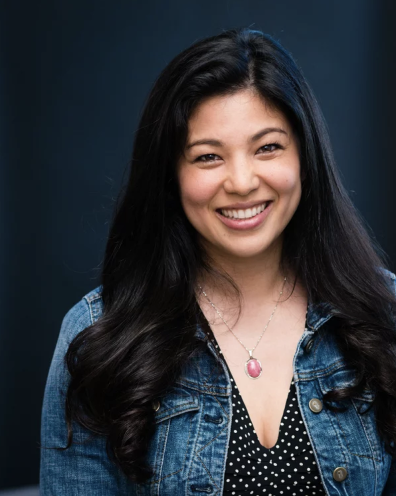 A young Asian-American woman wearing a spotted dress and denim jacket smiles warmly. She had long black hair.