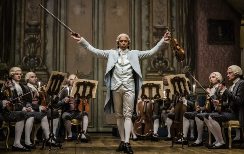A Black man in a fine period costume fitting of Marie Antoinette's time stands on stage, proudly holding aloft, in one hand a violin, in the other a bow. Seated musicians are lined up on both sides of him.