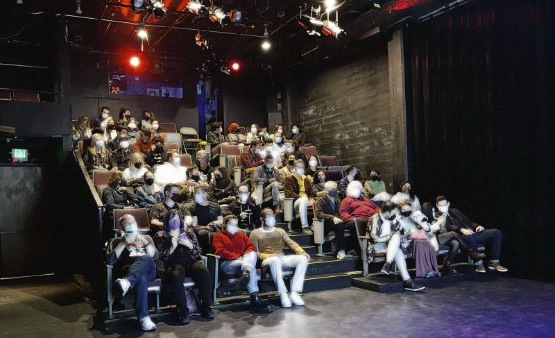 an audience of about 30 people, most of them wearing masks, is seen sitting in chairs watching a theatre performance