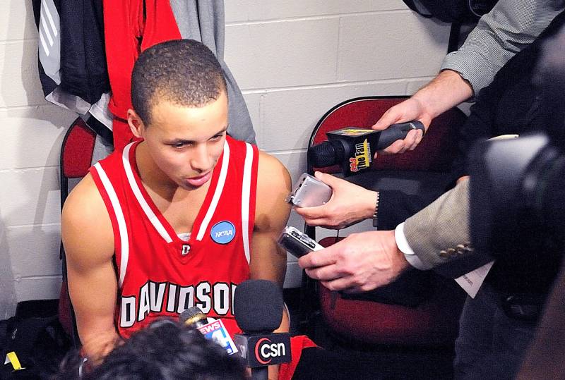 A college-aged Black man sits in the locker room with reporter microphones in his face