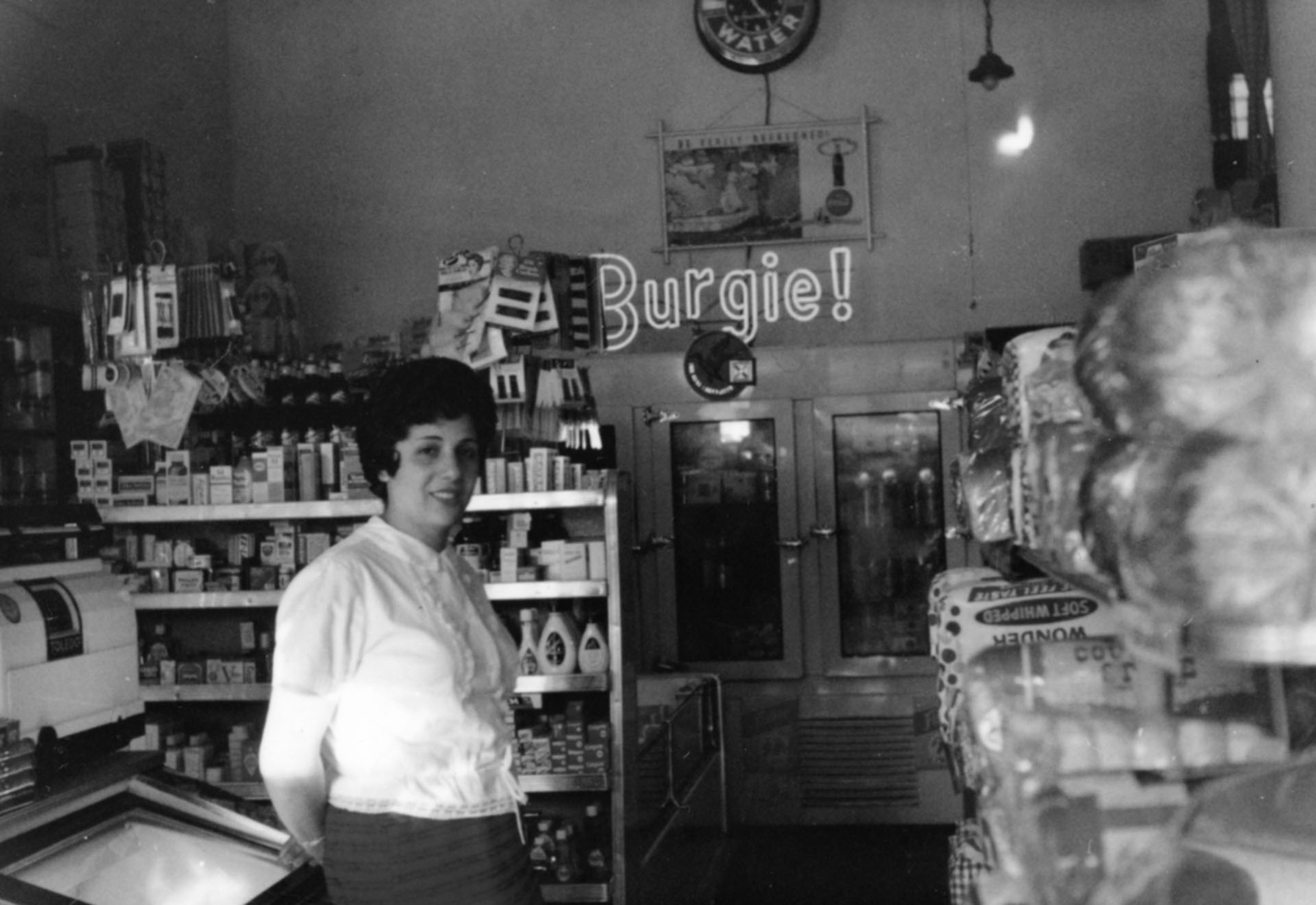 Old black-and-white photo of a woman standing inside a deli/market; a neon sign in back reads, "Burgie!"