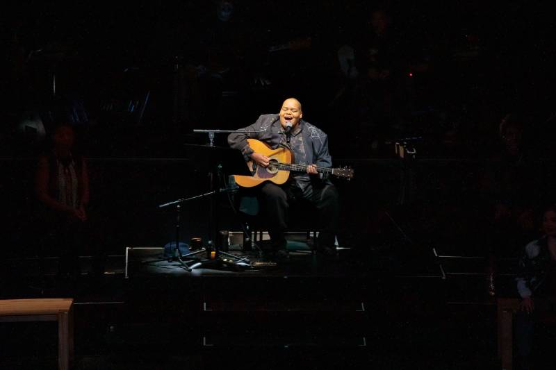Toshi Reagon plays her acoustic guitar on a dimly lit theater stage.