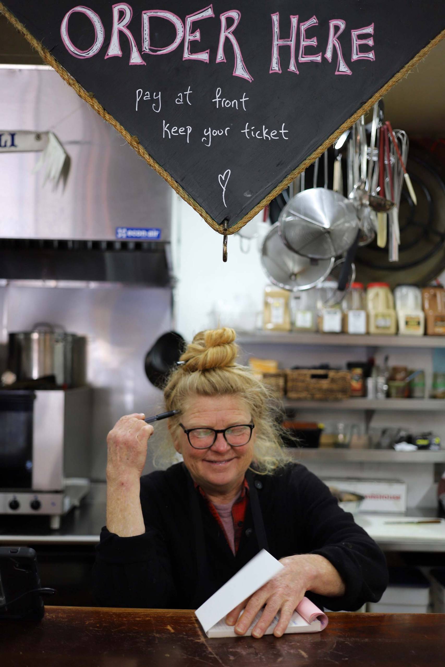 A middle-aged woman in glasses stands behind sa deli counter, her blonde hair tied in a bun.