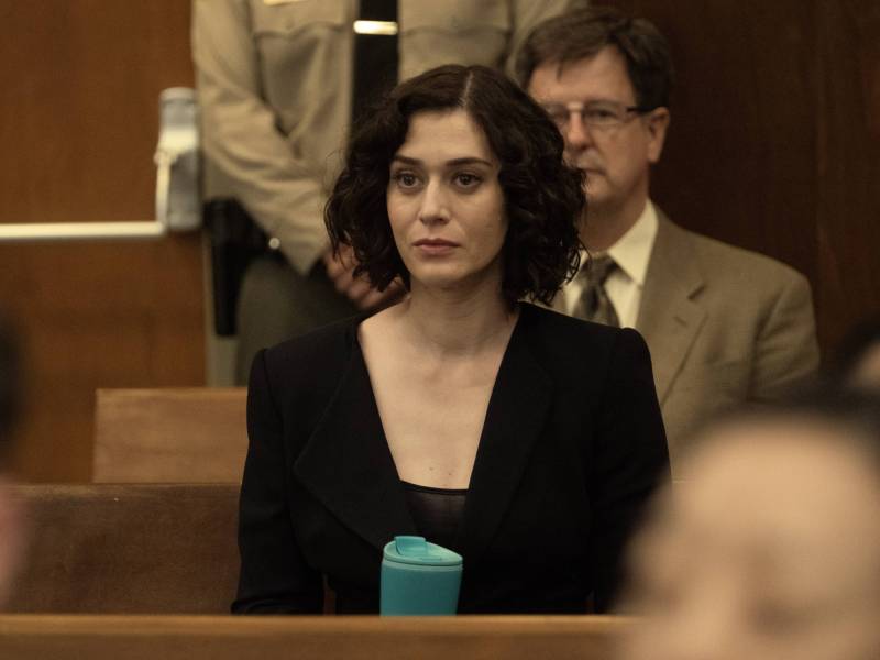 A pale woman sits in a courtroom, face impassive.