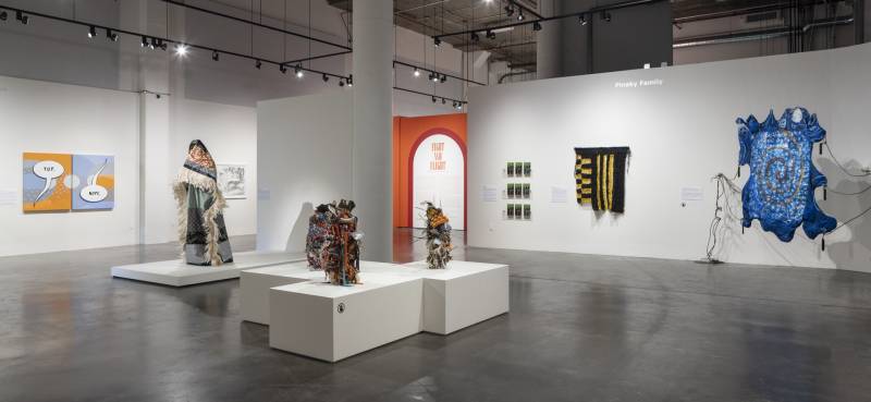 Wide photo of gallery install with colorful works on walls, sculptures on pedestals and exhibition title.