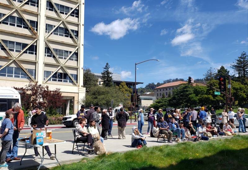 A crowd of people stand and sit in lawn chairs on a sunny street corner.