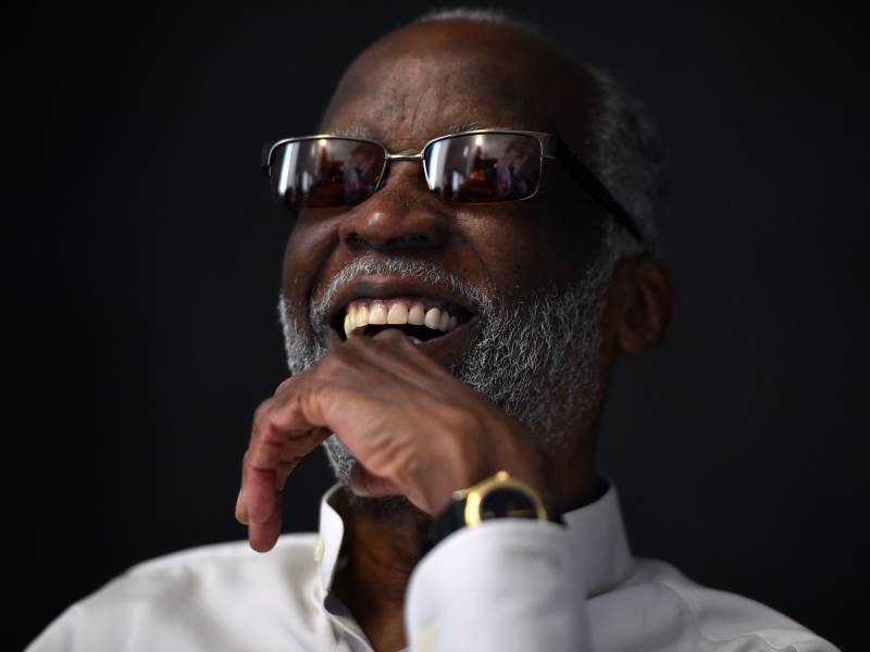A man in a white shirt, sunglasses and watch smiles, his head tilting backward