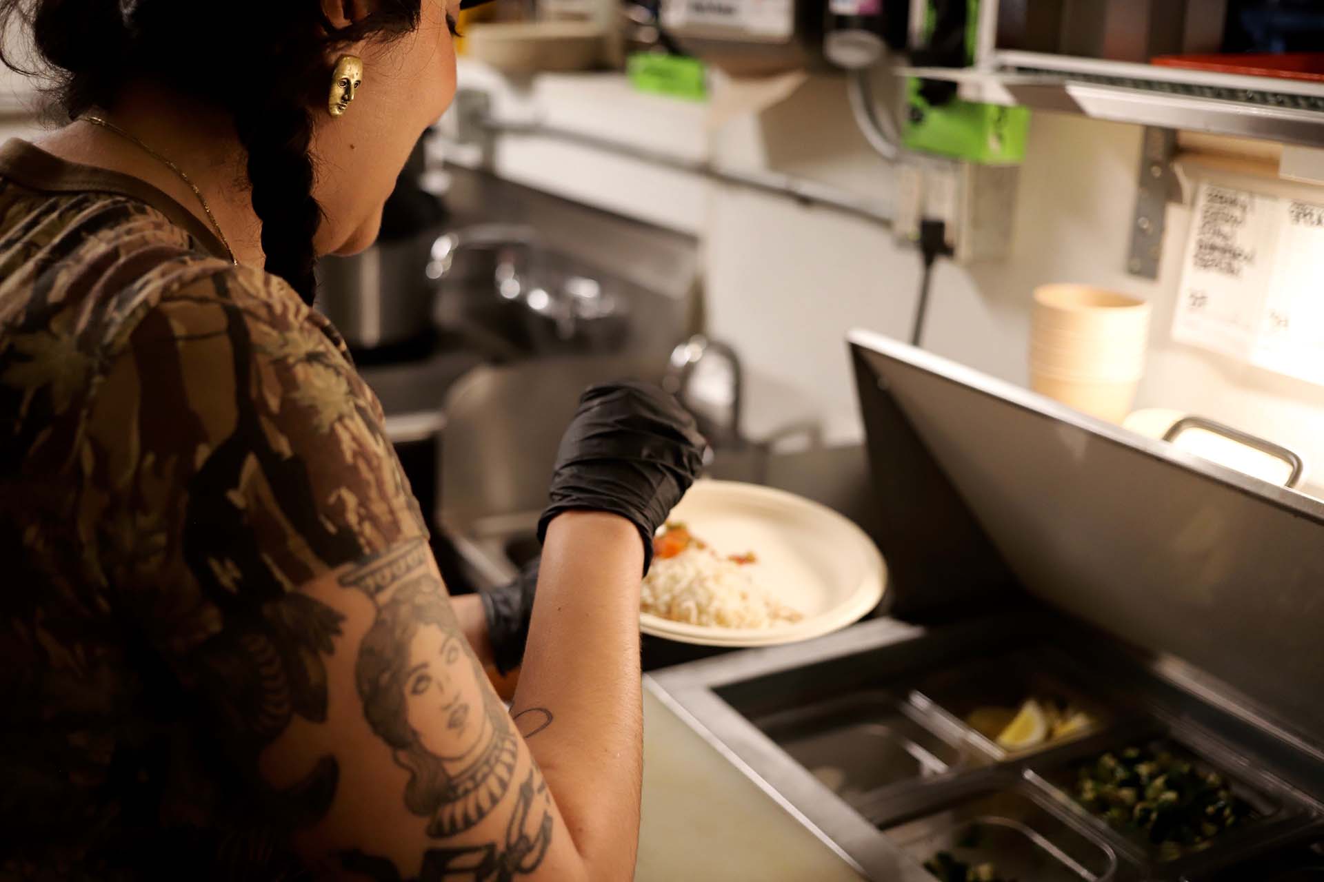 Tattooed woman wearing black plastic gloves prepares a plate of rice inside a restaurant kitchen.