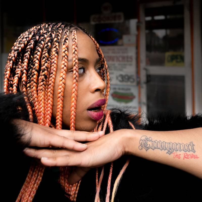 A young Black woman with bright extensions and an arm tattoo reading 'Cougnut - the Rebel' looks to the right with treated contacts