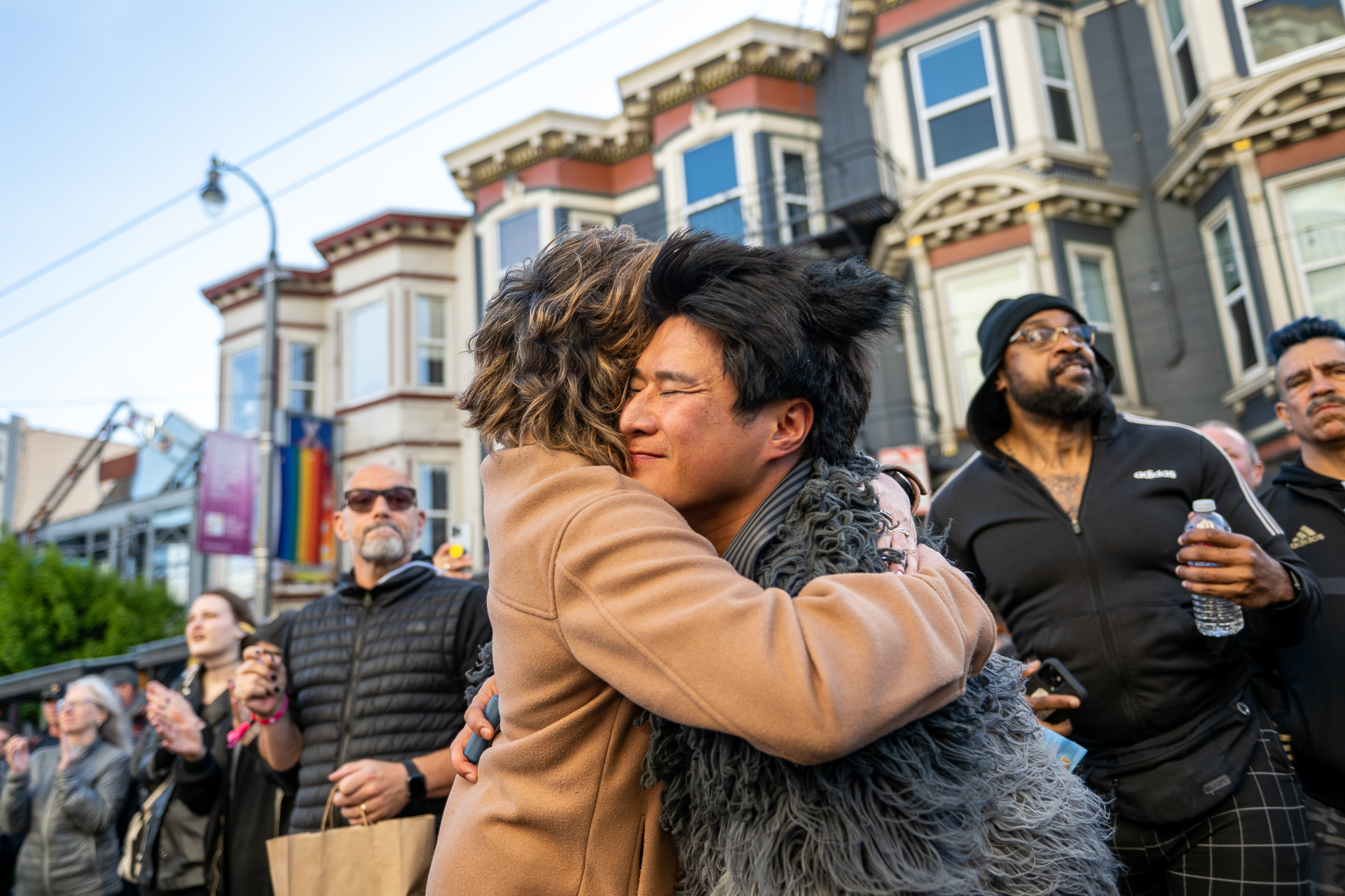 Two people share a deep hug and smile while they stand outdoors in San Francisco's Castro Street, surrounded by a large crowd.