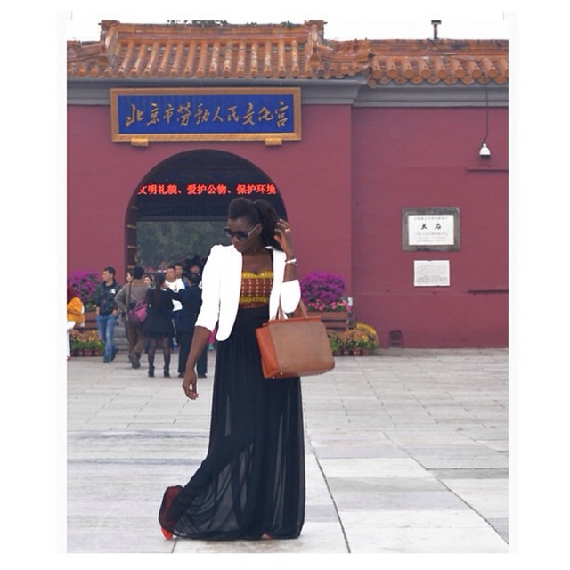 a photo of a slim Black woman in a dark dress and white jacket standing in front of a maroon building with Chinese lettering
