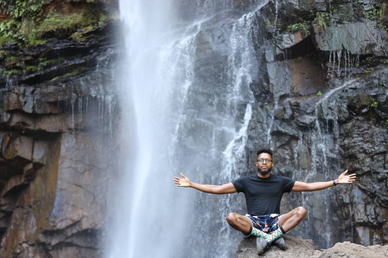 Anwar Bey sitting in the lotus position with his arms up while posing in front of a waterfall in South Africa.