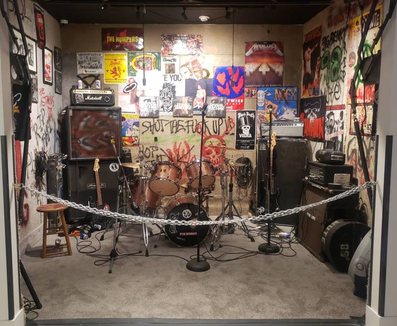A room with a security chain running in front of it contains musical equipment, a grimy carpet and numerous flyers and posters.