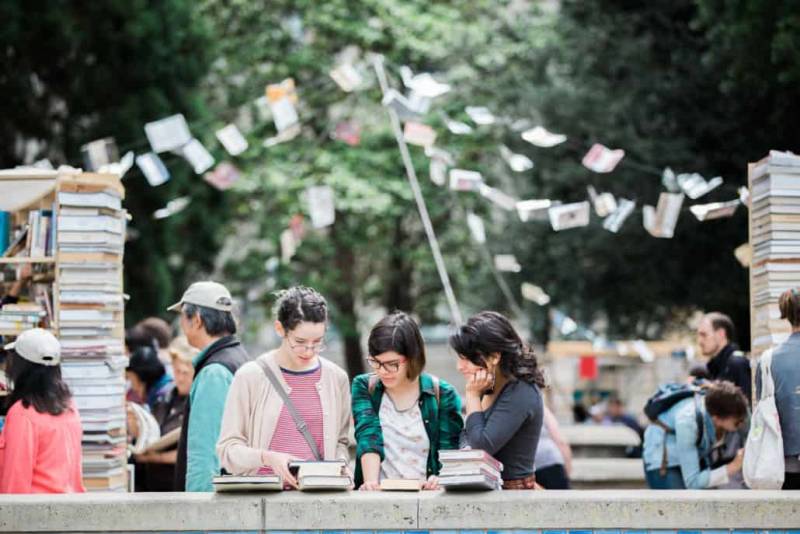 Three young women lean over their small stacks of books with banners and crowd behind