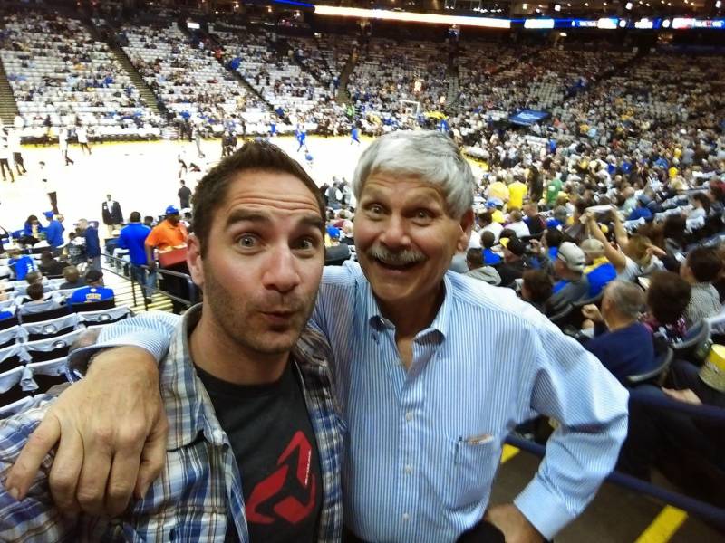 a young white man in a flannel shirt and an older man, his father, in a blue button up shirt, with their arms around each other, smiling for the camera at a basketball game